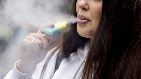 A close up of a young woman using a disposable vape. Only the bottom half of her face can be seen. She is wearing a white hoody. 