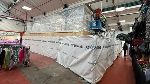 Construction work is starting in the marketplace, with an area protected by plastic sheeting and boards. A rail of clothes is standing to one side with a deli stall behind it. People are working on the site.