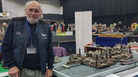 A man in his 80s dressed in navy stands beside a model of Mulberry Harbour