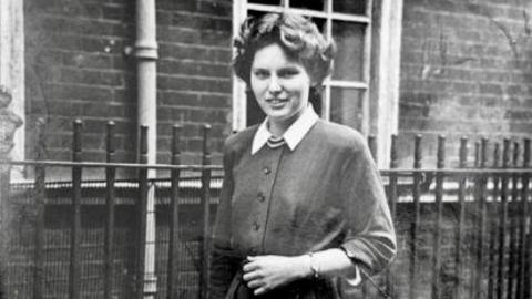 A black and white photo of a young woman with short dark curly hair. She wears a cardigan with white collar sleeves and stands in front of a brick building with a black fence.
