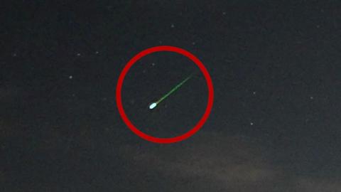 A meteor with a bright green tail streaks through the night sky, with several stars nearby. The meteor is circled with a red overlay.