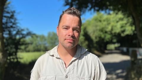 Stephen Le Quesne is stood on a Jersey lane wearing a khaki shirt.