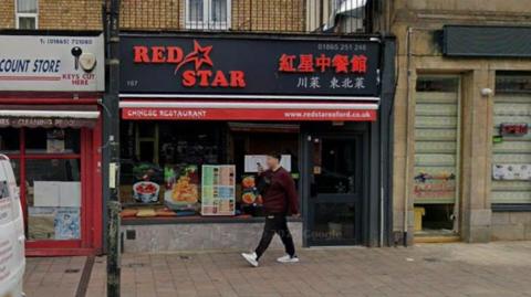 The front of the Chinese restaurant Red Star in Cowley Road. It's daytime and a man is walking past the restaurant while speaking into his phone. The restaurant's sign includes Chinese letters and a red star symbol. 