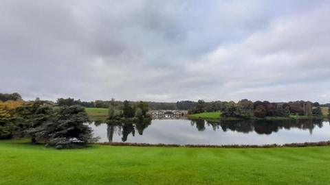 A large pool of water sits in the centre of the picture with trees standing either side. The water is completely still and you can see the trees reflecting in it. A bridge can be seen in the background