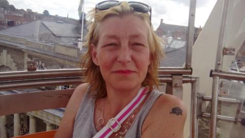 Dawn Sturgess looking at the camera. She has blonde hair and is wearing a grey vest top. She has sunglasses on her head and the strap of a bag over her shoulder is visible and is wearing a necklace. 