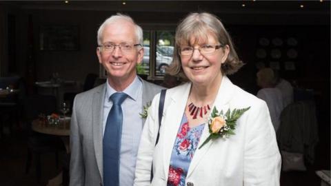 A couple smile at the camera. He is on the left and has short grey hair and glasses, and is wearing a grey suit with a blue short and tie. She is on the right, has short greying hair in a bob, glasses and is wearing a blue and red floral dress and a white jacket with a flower attached to the chest