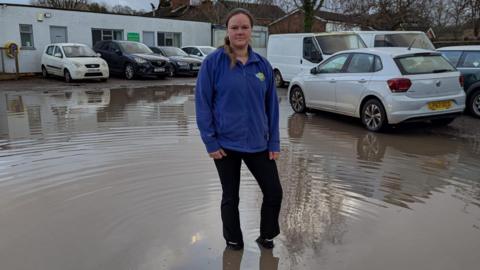 Woman in blue sweatshirt up to her ankles in water in a car park in front of a building