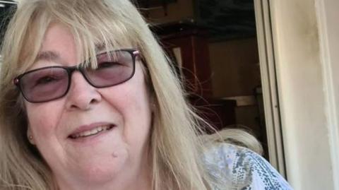 Elaine Lawrence, 71, who has shoulder-length blonde hair, is wearing tinted glasses and a blue and white blouse, smiles into the camera
