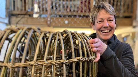 A smiling woman holding a lobster pot made of reeds.