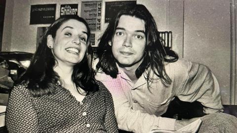 A black and white photo of Conal with his wife Lee. Conal has long dark hair and is wearing a shirt and his wife has long brown hair and a patterned button up blouse. They are both looking at the camera.