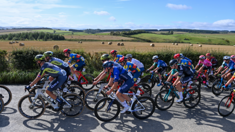 Several road cyclists dressed in various colours of lycra and helmets are all cycling down a country lane next to a field full of hay bales, with rolling fields behind it.