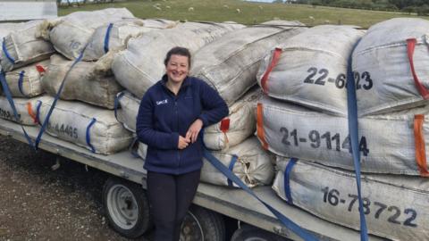 Emma Boocock stands in front of a truck of wool, packaged in sacks. She has brown hair tied up in a ponytail, and wears a navy blue fleece and black trousers. In the background, fields of sheep can be seen.