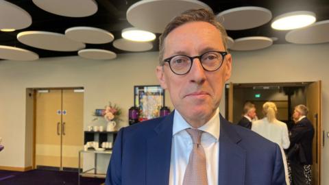 Deputy Ian Gorst is wearing a blue suit and white shirt with a pink tie. 