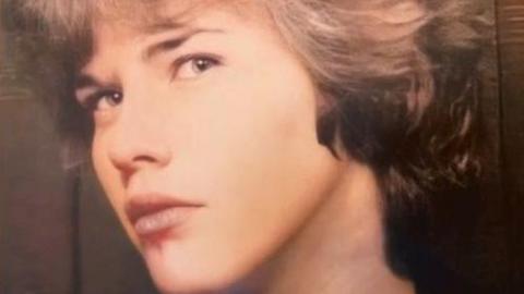 The picture is of a woman, looking side on to the camera, as if she is in a photo booth, as it is a close up of her face and he background is brown. She has very clear skin and short brown wavy hair. She is looking towards the camera and she is not smiling.