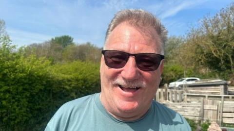 A man wearing sunglasses, a green T-shirt and an earring smiles at the camera.