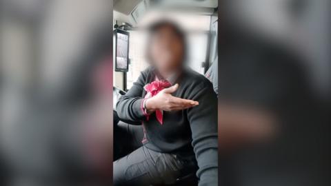 A bus driver gestures with one hand while wearing a black sweater and a red bow tie.