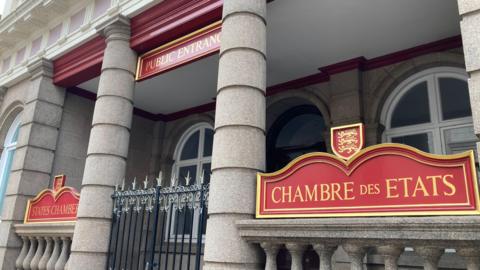 Jersey States Assembly building. The front of the building is made from stone and has red and gold signage. There is also a metal gate with spikes on it.