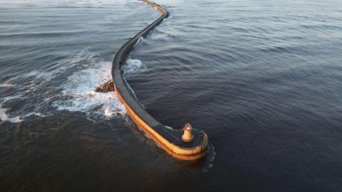 An aerial view of the lighthouse after its top was ripped off during Storm Babet. It shows the sunlit lighthouse with the pier in the distance in South Shields.