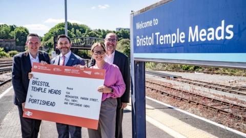 On a bright summer's day, from right to left, Bristol City Council leader Kevin Guy, West of England Mayor Helen Godwin, North Somerset MP Sadik Al Hassan and North Somerset Council leader Mike Bell all stand on a platform at Bristol Temple Meads, holding a giant train ticket. The ticket reads "from Bristol Temple Meads to Portishead" and notes the route as going via Bedminster, Parson Street, Pill and Portishead.