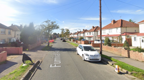 A Google street view screenshot of a sunny street with semi-detached houses on each side and cars parked on the roadside.