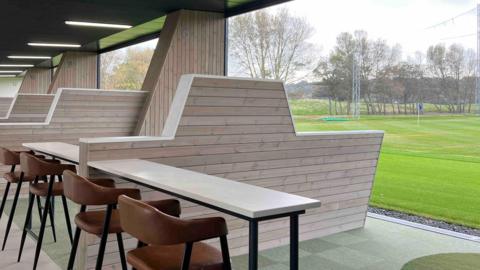 A golfing green is outside with a table and chars inside a driving range , with a large barrier between ranges under cover.