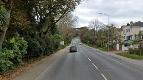 A picture of the street view in Falmouth. There is a large road with a few cars on it and houses to the right of the image. To the left, there is trees.