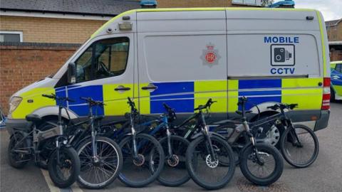 A mobile CCTV Gloucestershire Police van - which is mostly white with blue and yellow - with seven e-bikes lined up against it after being seized. There is a brick wall and a police car behind the van.