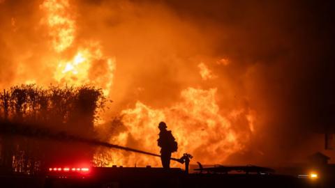 A firefighter and a hose spraying water are silhouetted against a wall of flames