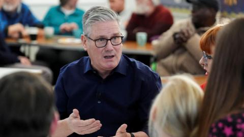 Sir Keir Starmer is wearing a navy shirt and has grey hair and place rimmed glasses. He is explaining something to a group of people who are out of focus on the edge of the image.  