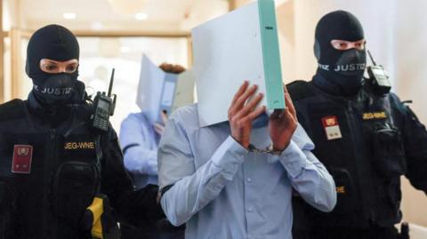 Two police officers in masks flank a suspect wearing a blue shirt and carrying a ring binder in front of his face