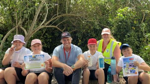 Charlotte, 10, Hattie, 9, Tony Tostevin (MUG), Ollie, 10, Juliet Bell (Vale Primary School Sun Safety Ambassador teacher) and Lewis, 9, sat in a shaded woodland area at the school. They are all smiling, wearing sun hats and holding an array of items like suncream, the sun-safe accreditation certificate and waterbotles. 