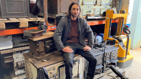 Olly Osborne in work clothes, sitting in his workshop on an old metal cooking range. There are a variety of other historic ovens to his side and behind him. He has long hair and a short beard. 