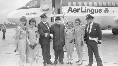 Black and white image of Monsignor James Horan stood stood in the middle of a group of pilots and air hostesses on an airport tarmac