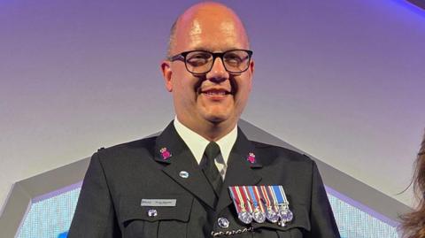 bald man wearing glasses and dark grey uniform with medals is smiling