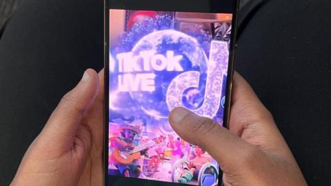 Shows someone's hands holding a mobile phone with TikTok Live on the screen.