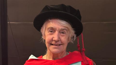 Lesley Gracie smiling at the camera wearing a black cap and red gown.