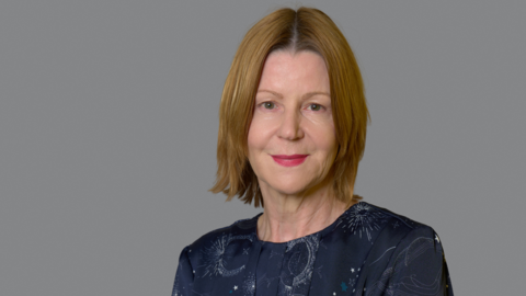 Maria Kane with short blonde hair and red lipstick looking at the camera and smiling. She is wearing a navy dress with moon and stars print, with a grey background. 