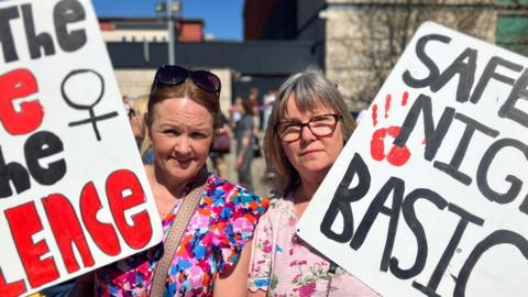 Two women are holding homemade posters. One of the women has auburn hair tied back and her sunglasses are perched on her head. She's wearing a colourful top. Her banner says: end the violence. The woman next to her wears tortoiseshell glasses and is wearing a pink floral top. Her banner says: safe. Other words are out of shot. People are standing in the background in the street. 
