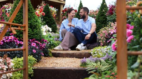 A couple pictured sitting on a step in a garden, laughing, surrounded by flowers.