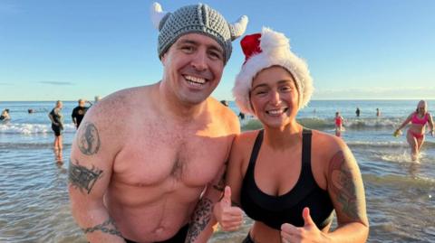 A man is wearing a knitted Viking hat and is bare chested. He has tattoos on both arms and is smiling broadly. He is standing next to a woman who is wearing a black bathing suit and Santa hat with her thumbs up. There are dippers in the waves behind them. 