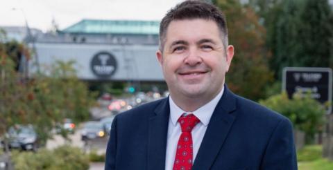 Shaun Davies MP is smiling at the camera, where a blue suit, white shirt and patterned red tie. Behind him is a road with traffic on, and commercial buildings beyond them