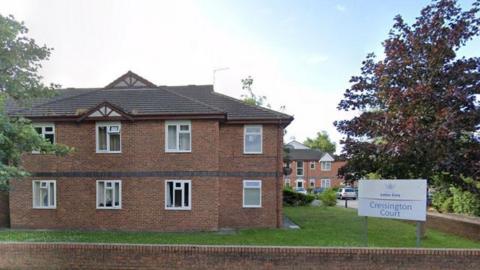 Cressington Court Care Home is a two-storey brick building surrounded by a lawn and trees. Its noticeboard is in the bottom right of the shot.