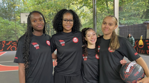 Four girls in black shirts and shorts stand together with their arms around each other and smile at the camera. They stand on a newly renovated outdoor basketball court.