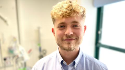 A young man with blonde hair and a beard, wearing a blue shirt.