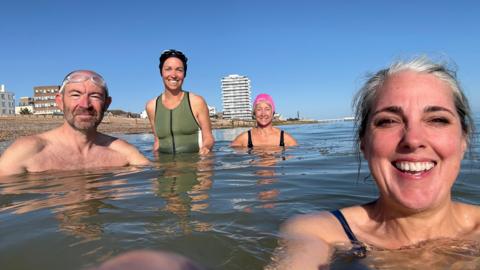 Three women and one man stood in the sea and smiling, not far from the shore. One woman has taking the picture, and the Worthing seafront can be seen in the background under a clear blue sky.