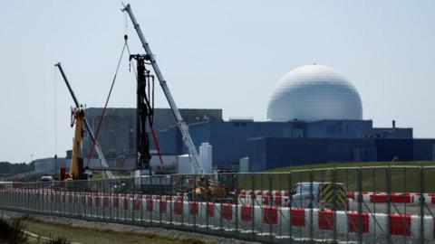 The large white dome of the Sizewell B nuclear power plant is the backdrop of the picture which has construction work and cranes in the foreground. The white dome sits on large square grey coloured buildings.