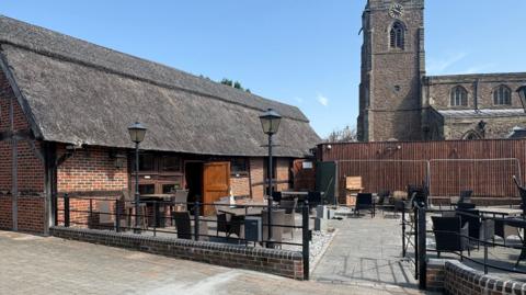 A cottage with a thatched roof and new fencing in front of church with spire