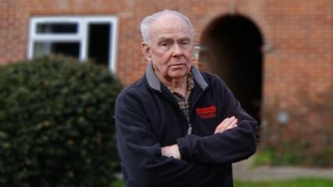 A man in a navy and/or grey fleece folding his arms in front of his terraced house. His white hair is receding.