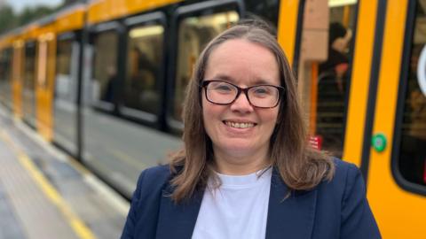 Cathy Massarella has black glasses and brown hair. She is wearing a blue blazer and a white top. She is smiling and standing in front of a new Tyne and Wear Metro train, which is out of focus. 