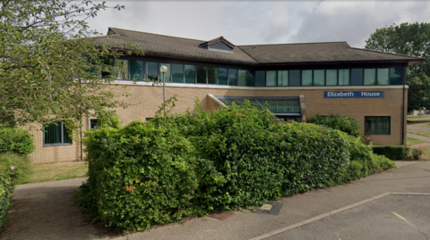 The outside of Elizabeth House, at Fulbourn Hospital. It is an angular brick building, with a line of windows on the top floor. In the foreground is a big green bush and in the distance, on the right, is a police car parked up.